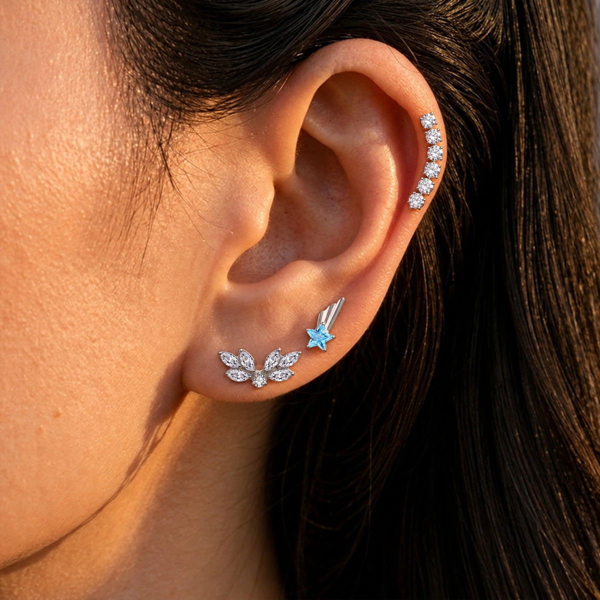 Close-up of an ear styled with a blue star-wing stud, a clear crystal angel-wing stud, and a curved crystal cartilage stud.