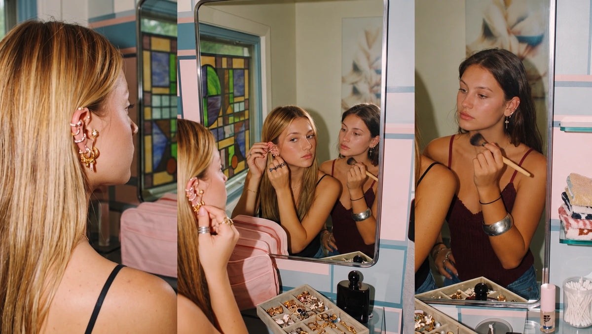 Two women styling stacked earrings and applying makeup in bathroom mirrors, with a jewelry tray on the counter.